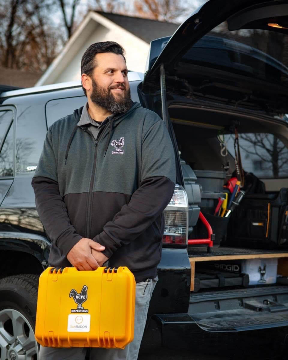 A home inspector wearing a custom branded jacket and holding a toolkit in front of a black truck.